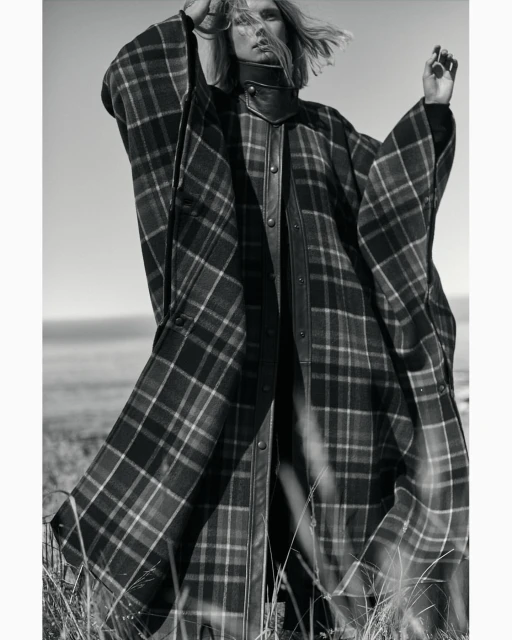 Monochrome portrait of a fair-skinned model in a plaid poncho-style coat. Tall grass is in the foreground, ocean and sky in the background.