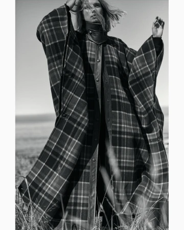 Monochrome portrait of a fair-skinned model in a plaid poncho-style coat. Tall grass is in the foreground, ocean and sky in the background.
