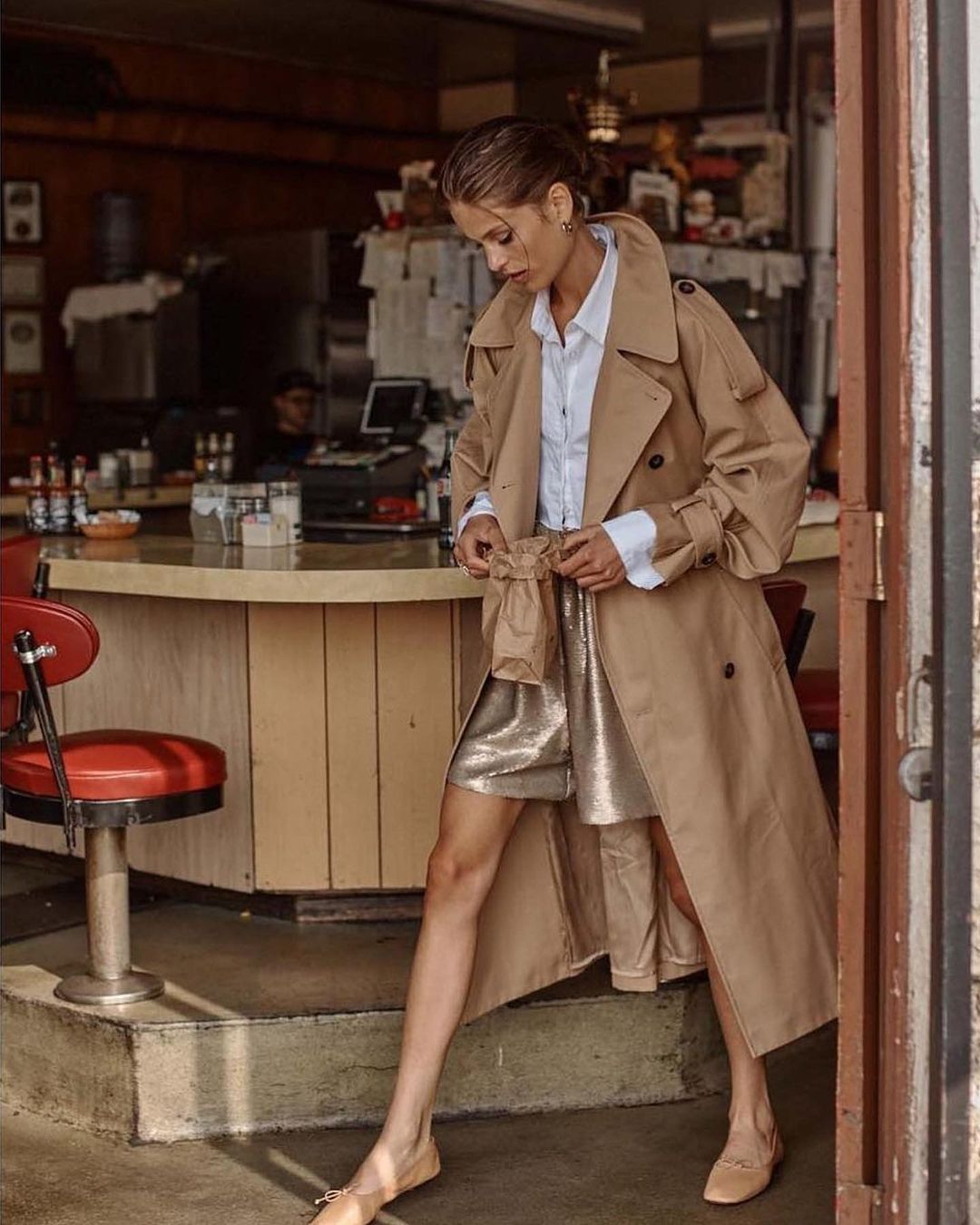 A stylish woman in a trench coat and gold sequin skirt poses in a vintage diner doorway, showcasing fashion and classic American style.