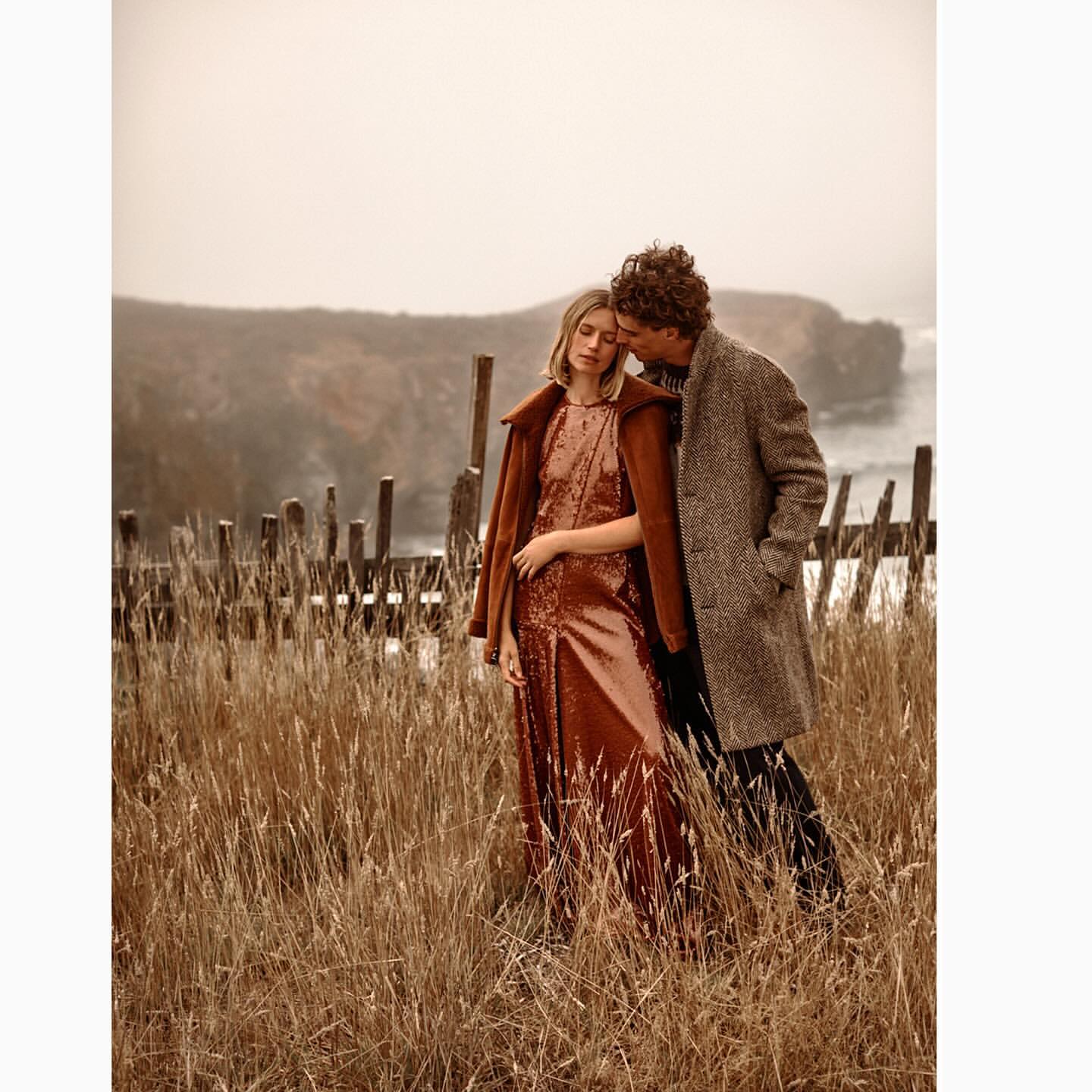 Fashionable couple in a field of wheat grass. The woman wears a sequined dress, the man a gray coat. Overcast sky and blurred cliff background.