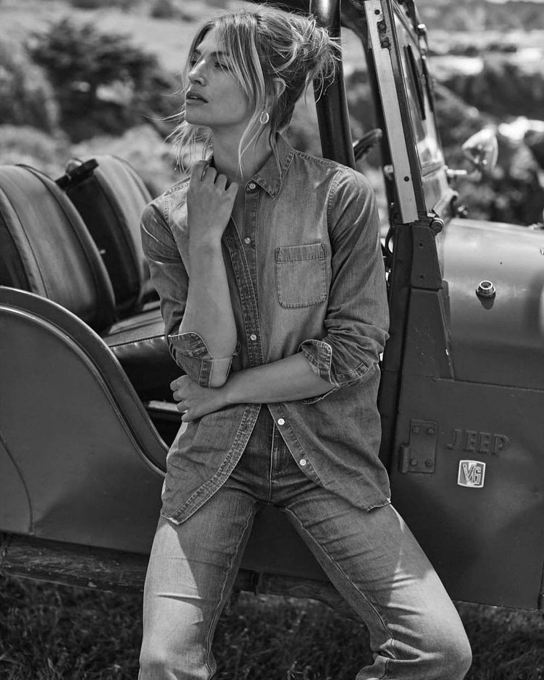 Black and white portrait of a young woman in a denim shirt and jeans sitting on the side of a Jeep in a grassy field.
