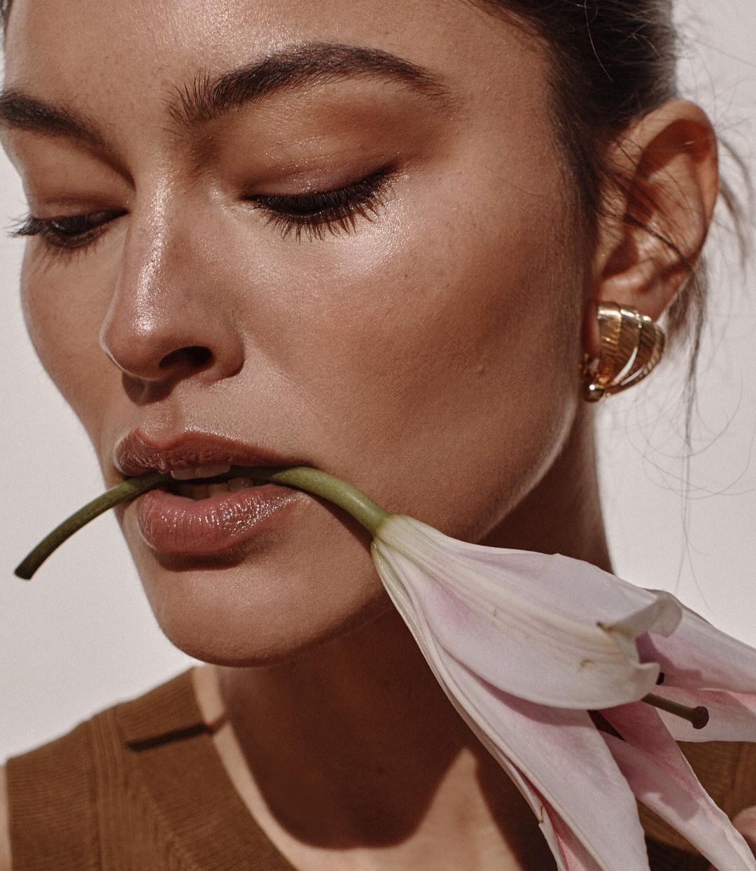 Close-up portrait of a fair-skinned woman with a flower stem in her mouth and pink lily, showcasing beauty and fashion.