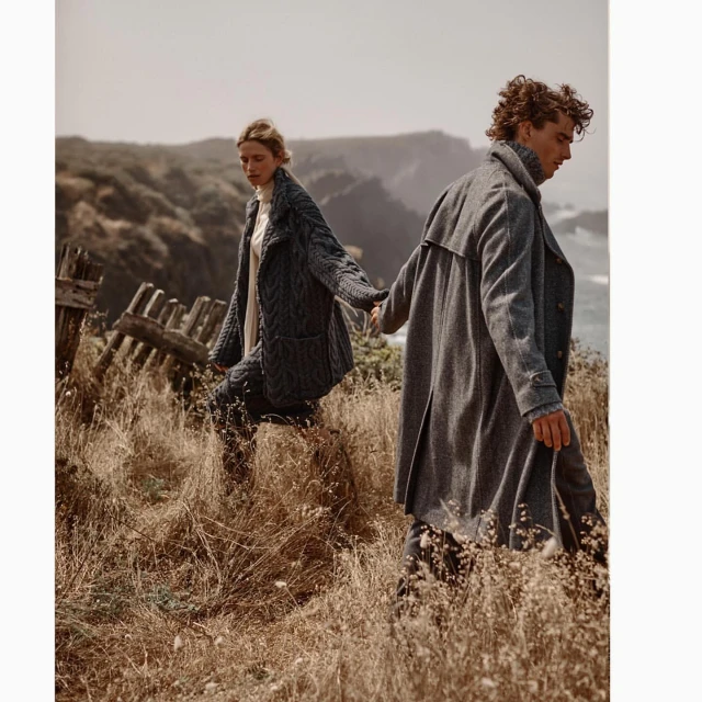 Couple in grey coats walking on a grassy hillside by the ocean; the woman wears a cable knit coat, the man a trench coat. 