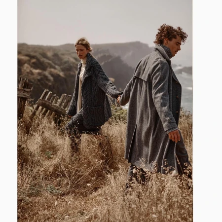 Couple in grey coats walking on a grassy hillside by the ocean; the woman wears a cable knit coat, the man a trench coat. 