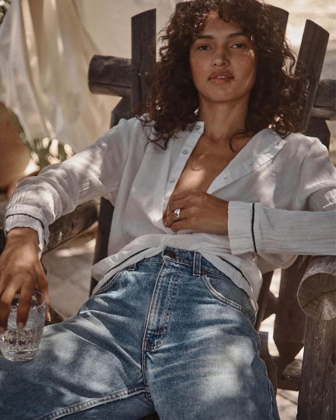 Brunette woman in a white shirt and jeans sitting in a wooden chair outdoors holding a glass of water in a lifestyle fashion shot.