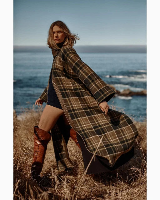 A woman in a plaid coat and thigh-high boots poses in a grassy coastal setting with the ocean in the background on a sunny day.