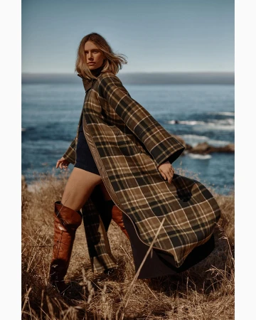 A woman in a plaid coat and thigh-high boots poses in a grassy coastal setting with the ocean in the background on a sunny day.