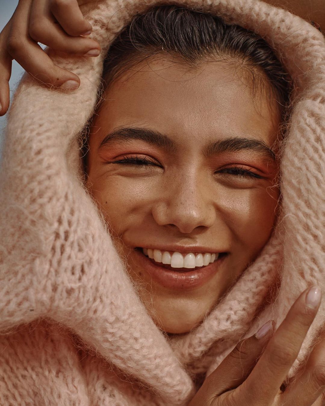 Close-up portrait of a smiling woman wrapped in a pink knit scarf, hands near her face, showcasing coral eyeshadow in an outdoor setting.