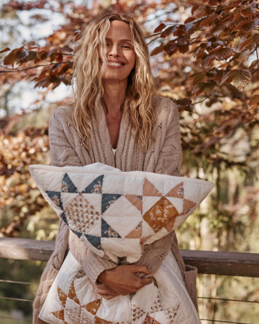 Smiling woman holding a star patterned quilt outdoors. She is wearing a cardigan and the background includes branches with brown leaves.