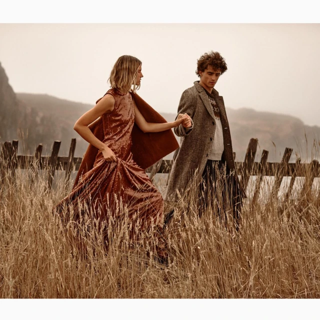 Woman in sequin dress and man in coat holding hands in a field of tall grass, with a hill and overcast sky in the background.