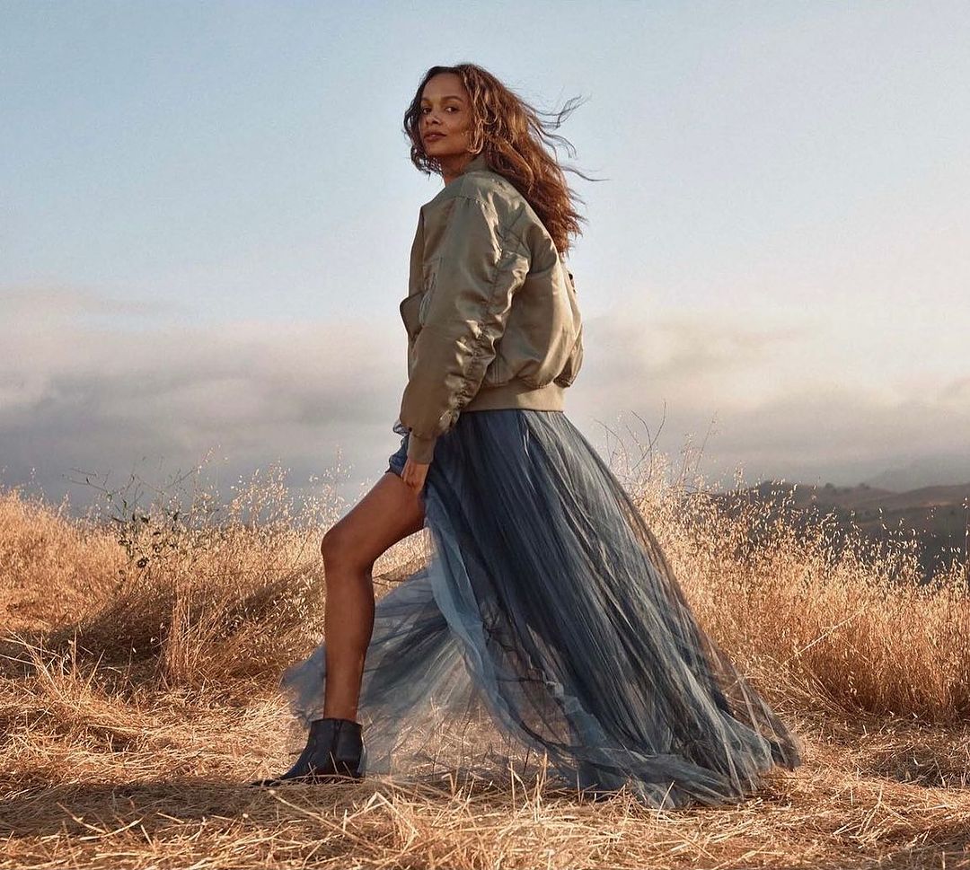A woman stands in a field of dry grass. She is wearing a khaki bomber jacket and a long blue tulle skirt with black boots. 
