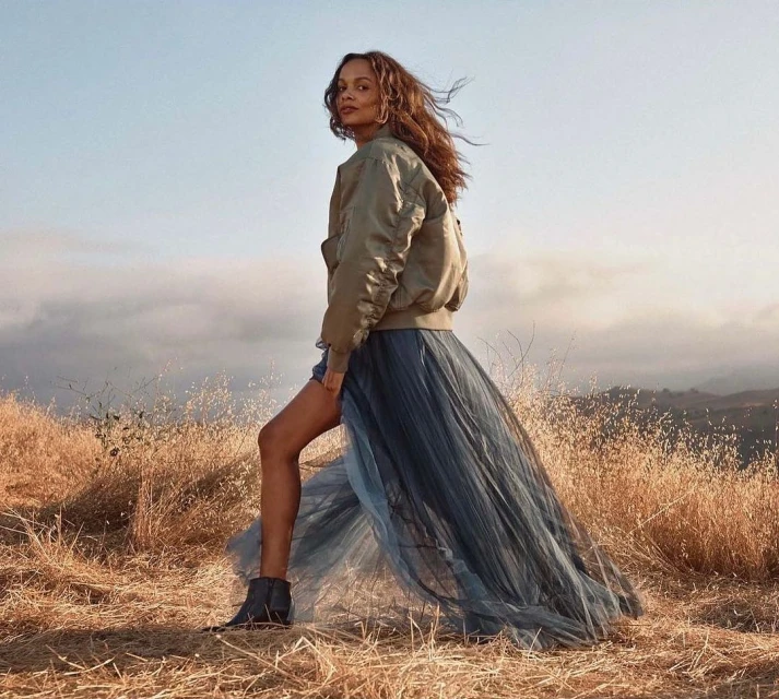 A woman stands in a field of dry grass. She is wearing a khaki bomber jacket and a long blue tulle skirt with black boots. 