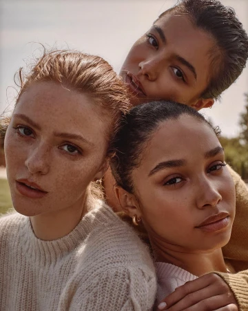 Close-up portrait of three young female models posing together outdoors in warm sunlight wearing knitted sweaters.