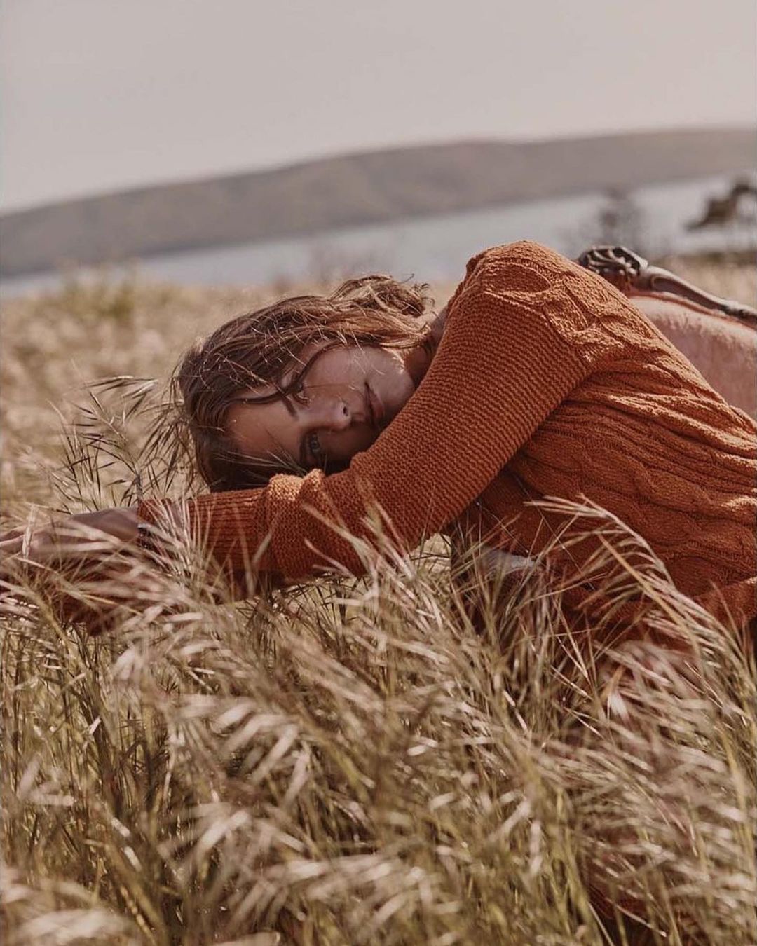 Woman in a rust-orange cable knit sweater lying in tall dry grass outdoors under an overcast sky, captured by Colette De Barros.