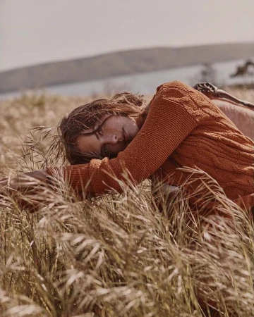 Woman in a rust-orange cable knit sweater lying in tall dry grass outdoors under an overcast sky, captured by Colette De Barros.
