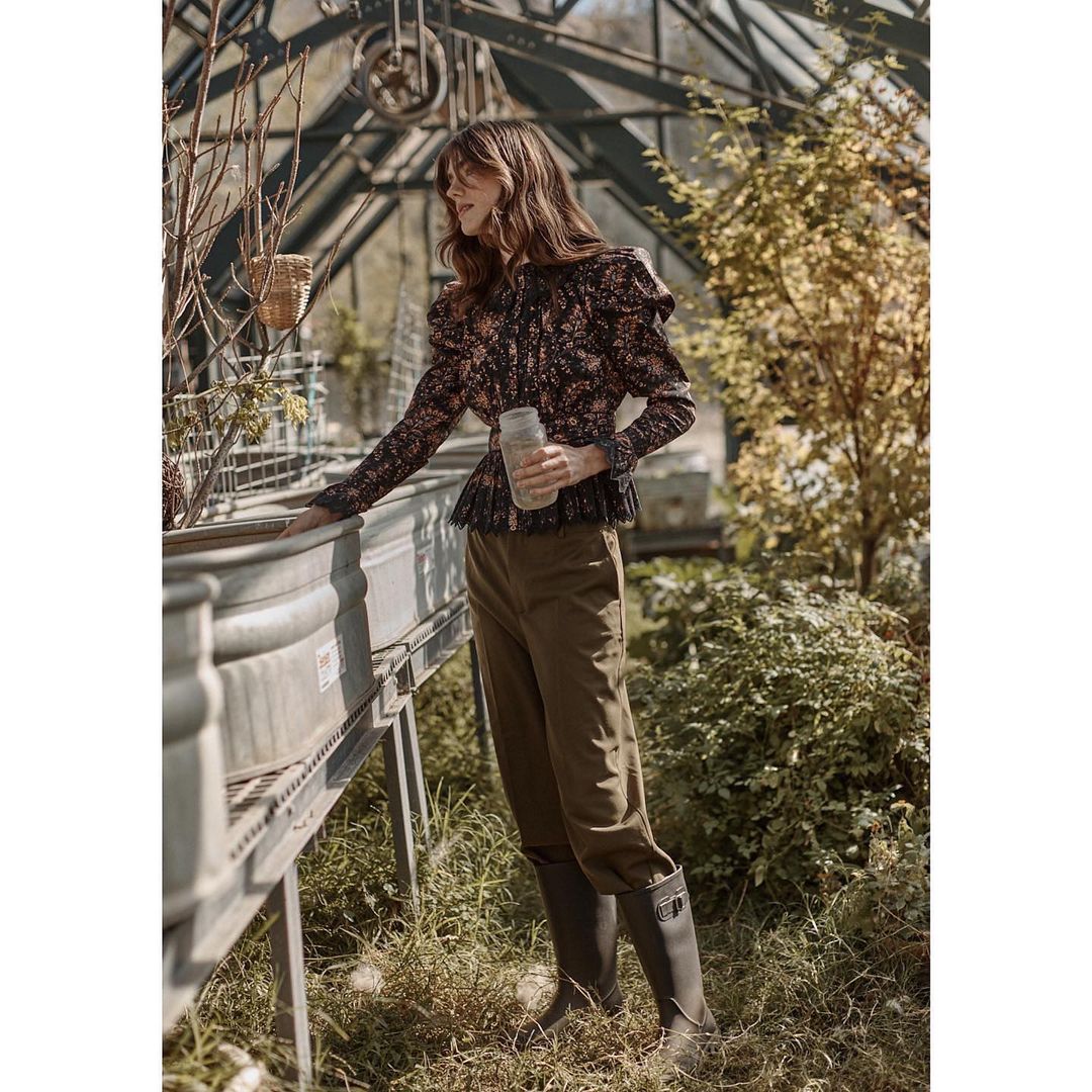 Adult woman in a floral shirt, dark pants, and black boots stands in a greenhouse surrounded by plants and gardening equipment.