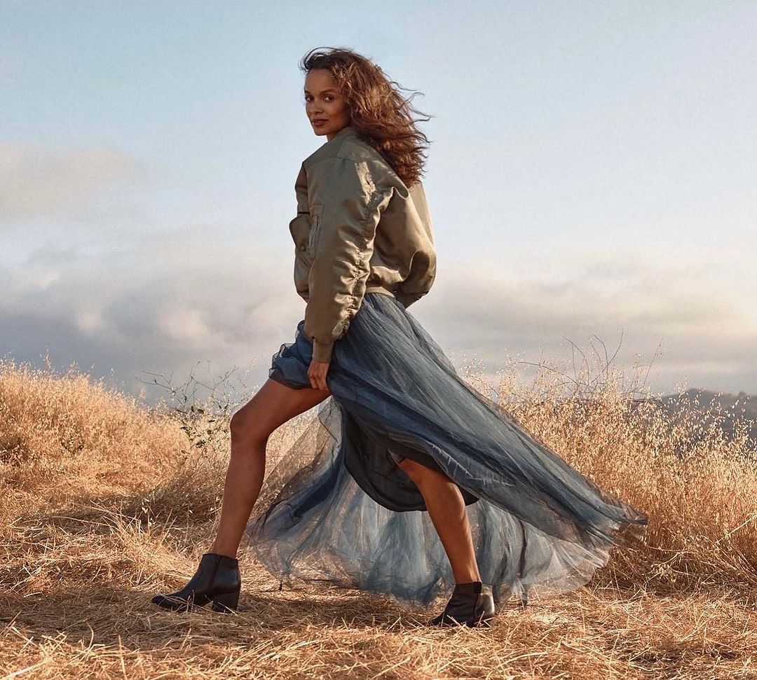 A woman with curly hair stands in a field wearing an olive bomber jacket and blue tulle skirt, posing while looking back over her shoulder.