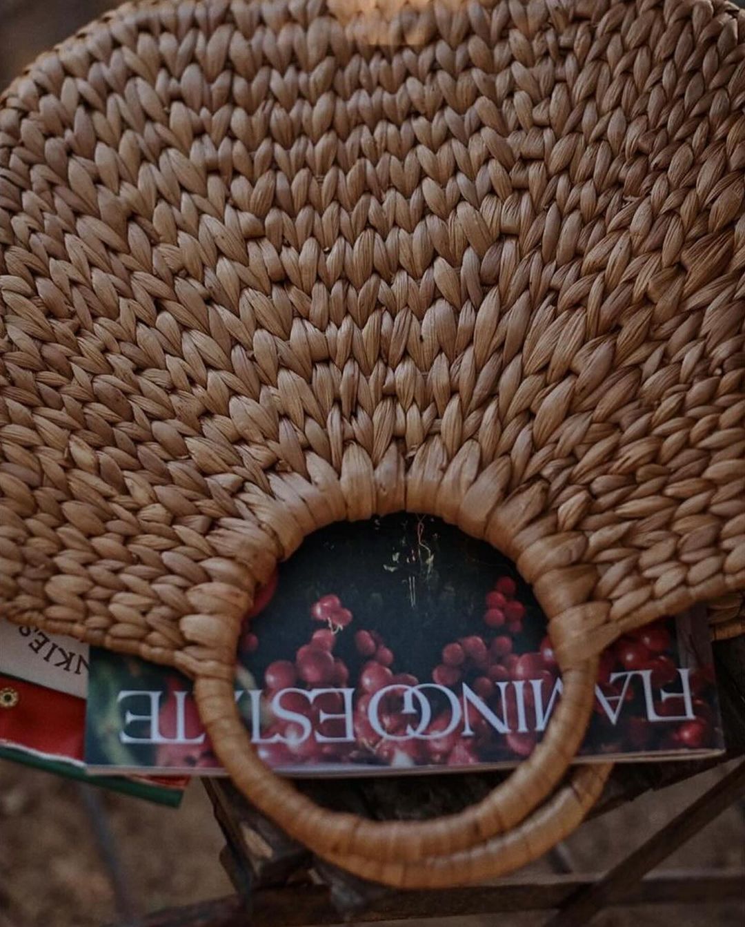 Close-up of a round, woven straw tote bag with books inside, sitting on a dark metal surface. Natural light gives a warm glow.