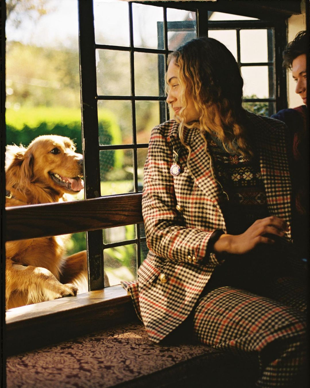 Woman in plaid suit sitting inside by a window with a Golden Retriever dog outdoors, looking into the house, with natural light.
