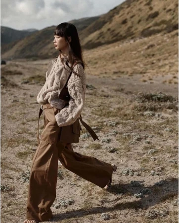 Model in chunky cable-knit sweater and brown trousers walking across barren landscape by Colette De Barros