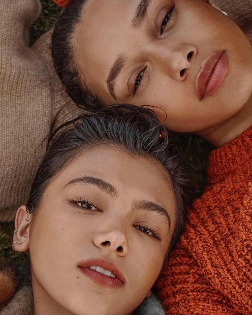 Two young women lie side-by-side outside, wearing fall-colored knit sweaters and natural makeup in a portrait shot, close to the camera.
