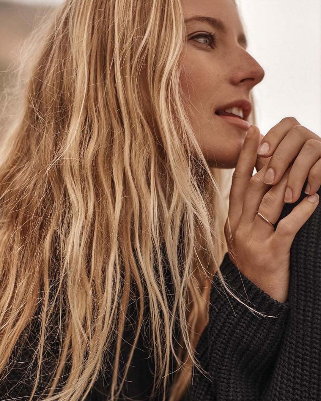 Close-up of a young adult woman with long blonde hair wearing a dark sweater and a ring on her finger, neutral outdoor background.