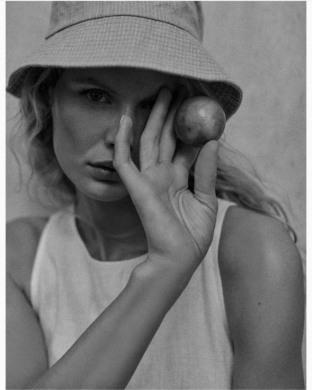 Monochrome portrait of a young woman wearing a bucket hat and sleeveless shirt, holding a round fruit near her face in a studio setting.