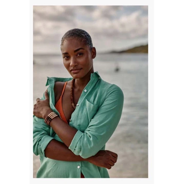 Portrait of a smiling woman in a teal shirt on a beach with a necklace, bracelet, and orange top on a cloudy day.