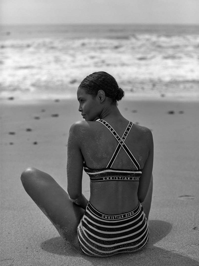 Black and white photo of a woman seated on a sandy beach wearing a striped bikini with branded straps, looking back toward the ocean.