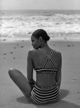 Black and white photo of a woman seated on a sandy beach wearing a striped bikini with branded straps, looking back toward the ocean.
