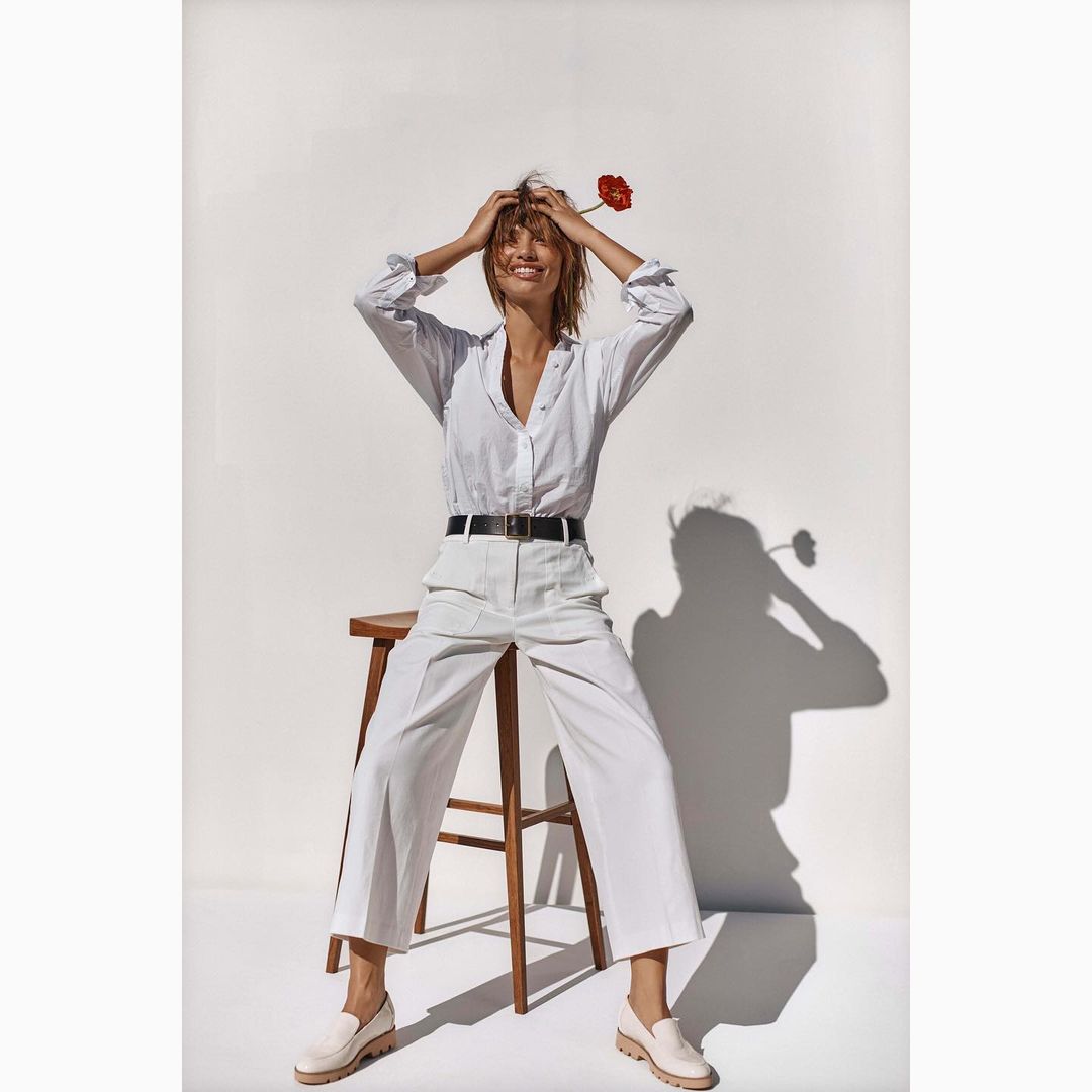 Woman with flower poses in white button-down shirt and trousers sitting on a stool in a bright indoor photo studio.