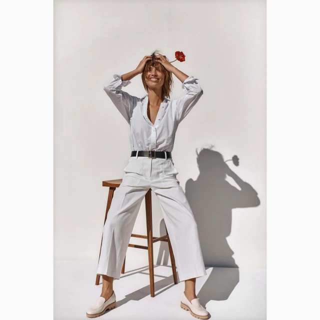 Woman with flower poses in white button-down shirt and trousers sitting on a stool in a bright indoor photo studio.