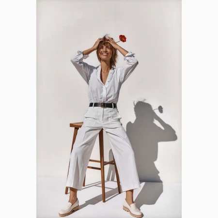 Woman with flower poses in white button-down shirt and trousers sitting on a stool in a bright indoor photo studio.