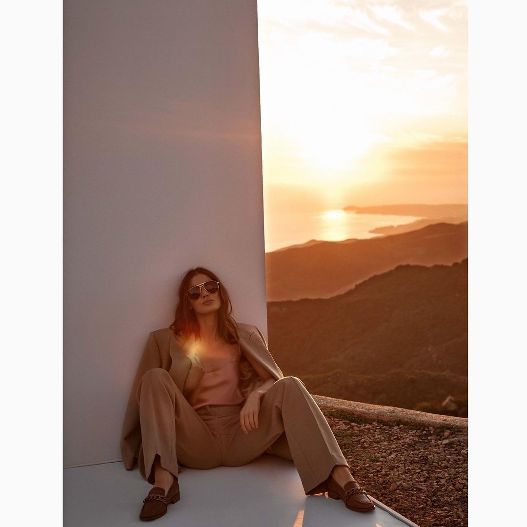Adult female in a tan business suit and sunglasses poses outdoors on a ledge overlooking mountains, a body of water, and a sunrise.