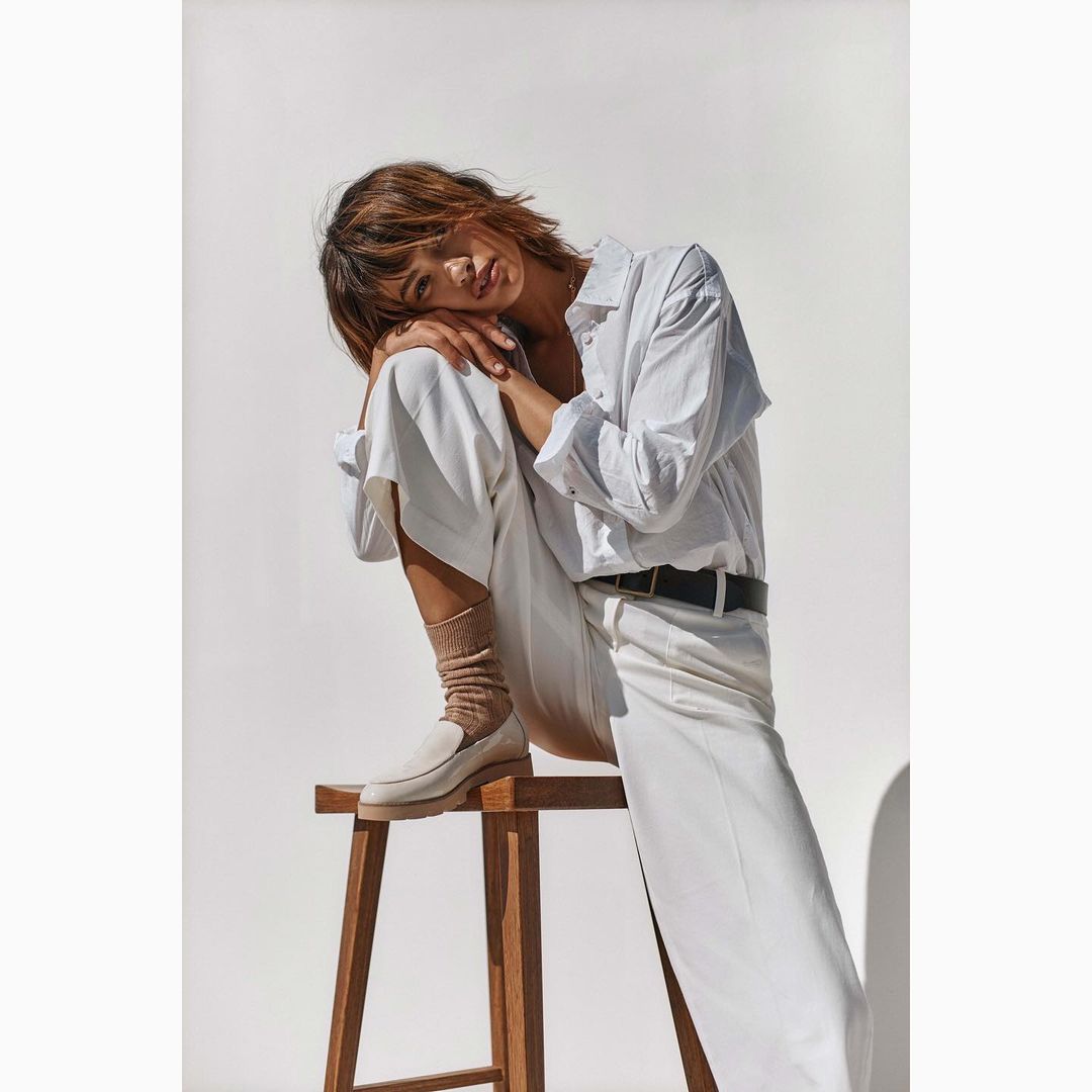 Woman in white shirt and pants sits on a wooden stool against a white background, styled with neutral socks and loafers.