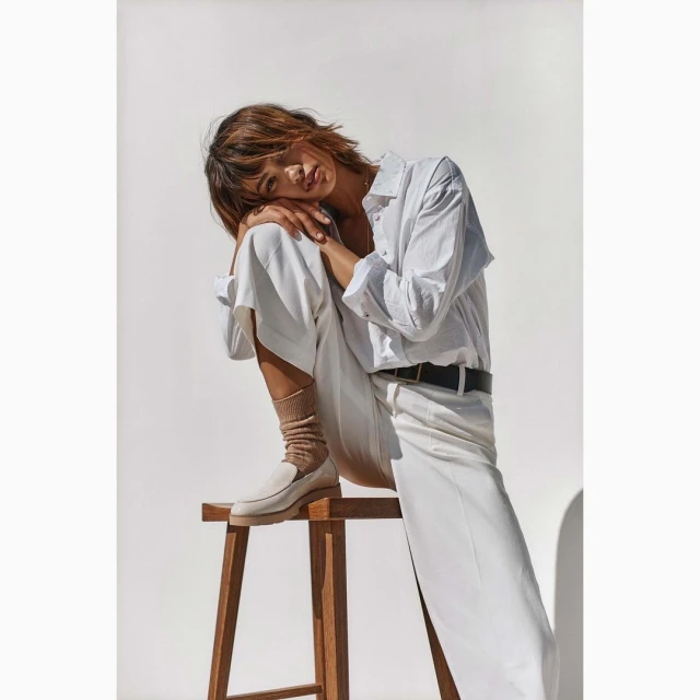 Woman in white shirt and pants sits on a wooden stool against a white background, styled with neutral socks and loafers.