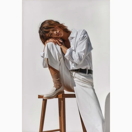Woman in white shirt and pants sits on a wooden stool against a white background, styled with neutral socks and loafers.