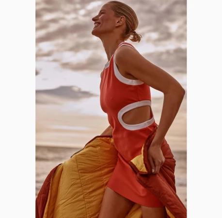 A woman in an orange dress smiles against a sky backdrop, holding a yellow and burgundy jacket in a beach setting during daytime.