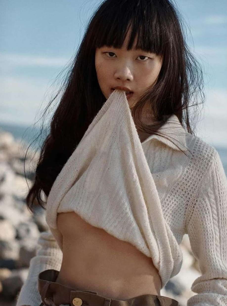 Close-up portrait of a woman lifting her cream cable-knit sweater near a rocky coastline by Colette De Barros