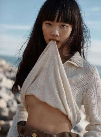Close-up portrait of a woman lifting her cream cable-knit sweater near a rocky coastline by Colette De Barros