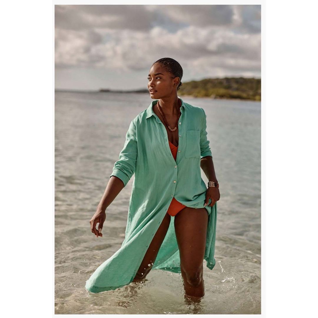 Woman in a long green shirt and swimsuit standing in ocean water on a sunny day, looking away from the camera, outdoor setting.