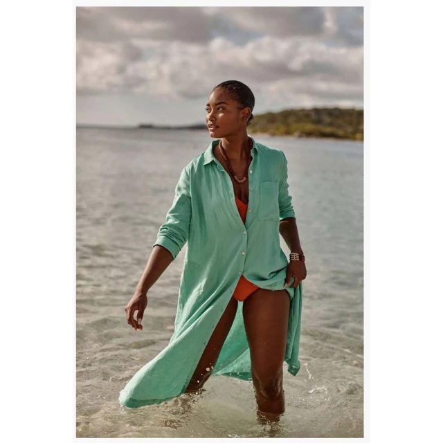 Woman in a long green shirt and swimsuit standing in ocean water on a sunny day, looking away from the camera, outdoor setting.
