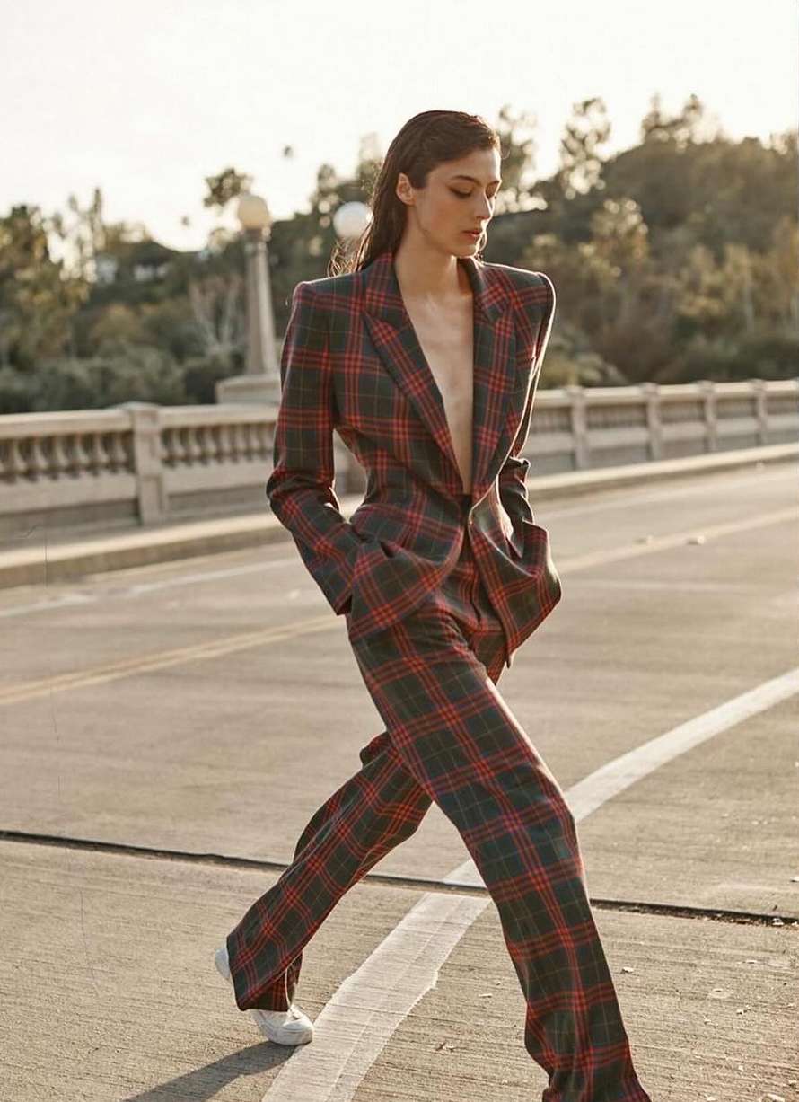 Woman in a plaid suit walks on an empty street. The model wears an open jacket and matching pants in daylight.