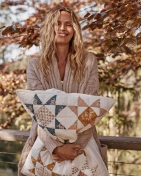 Woman with long blonde hair smiling while holding a patterned quilted pillow outdoors near trees.