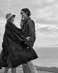 Black and white portrait of a smiling couple outdoors near the ocean holding an umbrella and wearing coats.