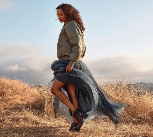 Woman in an olive green bomber jacket and layered dark tulle skirt walking through dry grassland at sunset