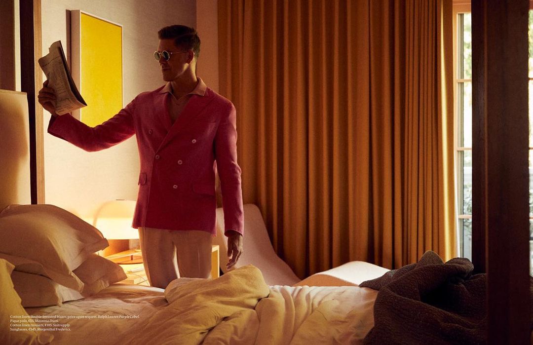 Man in a pink blazer reads a newspaper in a brightly lit hotel room with a bed, gold curtains, and a yellow painting on the wall.