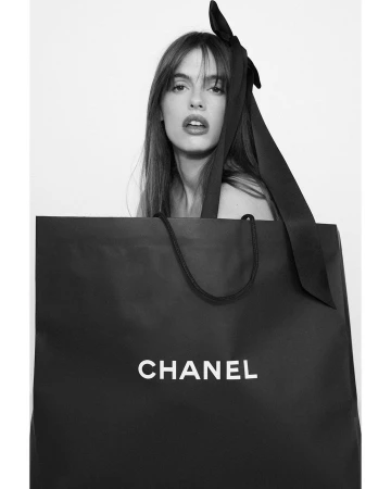 A woman peeking over a large black Chanel shopping bag with a long black hair ribbon in her hair