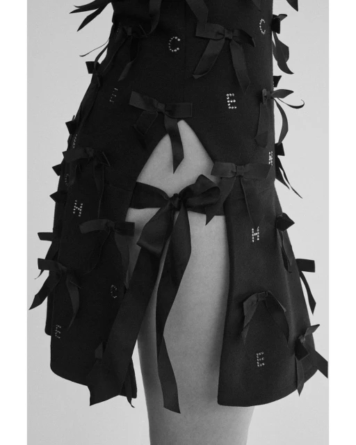 Close-up of a black fashion dress adorned with bows and letter embellishments, set against a plain white background in a studio.