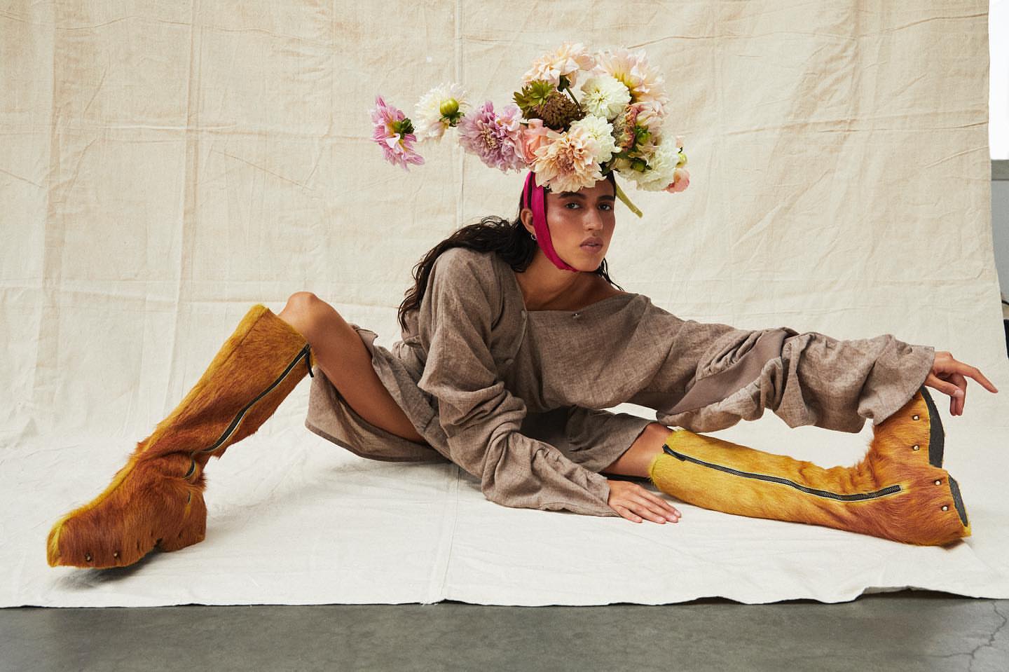 A woman in a grey dress sits on a white backdrop wearing tall fur boots and a large floral headpiece in a studio setting.